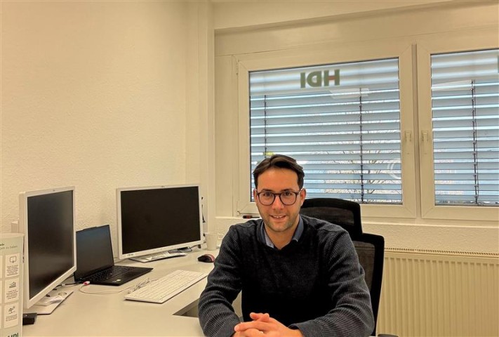 Akcan sitzt an seinem Schreibtisch und guckt in die Kamera. Links hinter ihm geht der Schreibtisch weiter und dort sind zwei Bildschirme, ein Laptop und eine Tastatur.