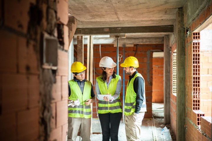 Eine Frau, in der Mitte und zwei Männe, links und rechts neben der Frau. Sie tragen alle eine Schutzweste und einen Helm, außerdem gucken alle auf den Plan, den die Frau in der Hand hält. Sie stehen auf einer Baustelle.