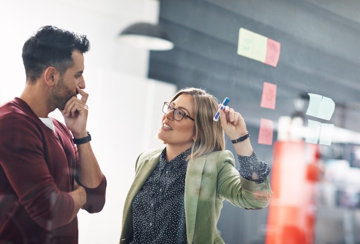 Links steht ein Mitarbeiter und Rechts ist eine Mitarbeiterin, die einen blauen Stift in der linken Hand hält, die nach oben gerichtet ist. Bei der Unterhaltungen gucken sich beide an.
