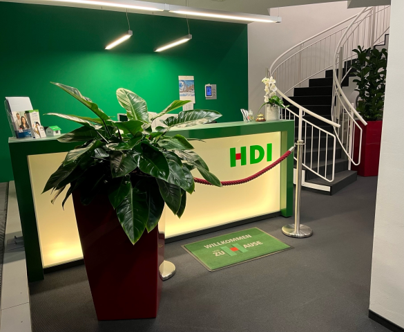 Tresen mit dem HDI Logo. Rechts daneben führt ein geschwungene Treppe ins Obergeschoss. Vorne rechts im Bild steht eine große Zimmerpflanze.