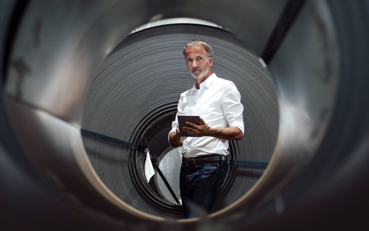 Mann mit weißem Hemd und grauem Bart steht mit einem Tablet in der Hand vor einer schwarzen Röhre
