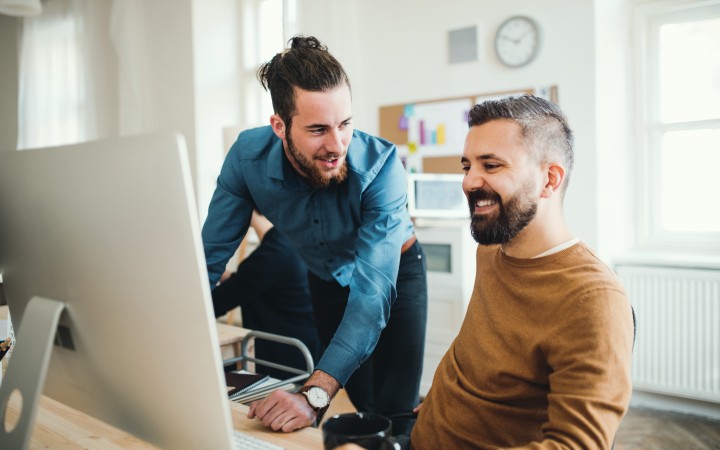 Zwei Männer an Schreibtisch, der Mann erklärt dem anderen etwas am PC