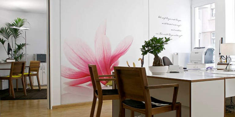 Büro mit einem Tisch und Stühlen und einer weißen Tapete auf der eine rosa Blume ist. Links ist ein Durchgang ,der in ein weiteres Büro führt, indem ein Tisch, zwei Stühle, eine Lampe und eine Pflanze steht.