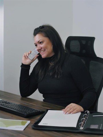 Frau Nebuert sitzt am Schreibtisch, vor liegt eine Tastatur und ein schwarzer Ordner. In ihrer linken Hand hält sie sich ein Telefon an ihr Ohr, dabei lächelt sie.