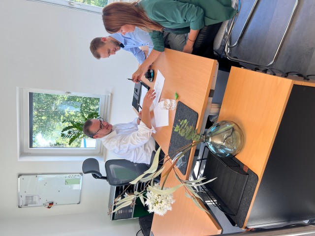 Ein Tisch steht in der Mitte, links vom Tisch sitzt Agenturleiter Keil und zeigt etwas auf einem Zettel. Ihm gegenüber sitzt eine Frau und ein Mann, die Frau hält einen Stift in der Hand. 