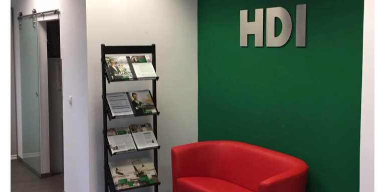 Ein roter Couch steht vor einer grünen Wand, an der in silber das HDI Logo hängt. Links neben der Couch steht ein aufsteller mit Zeitschriften.