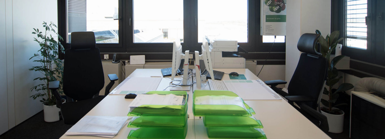 Links steht ein weißer Schreibtisch mit einem PC und rechts ist ein schwarzer Stuhl. Im Hintergrund ist ein Fenster.