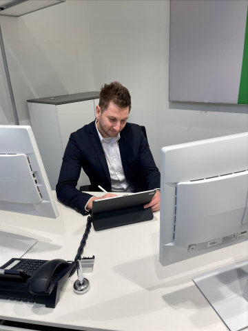 Mitarbeiter Sven Mayer sietzt an einem Schreibtisch, vor zwei Bildschirmen und guckt nach unten auf ein Ipad und schreibt etwas darauf. Hinter Ihm steht ein weißes Regal.