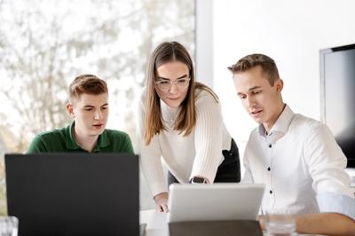 Drei Mitarbeiter stehen und sitzen an einem Tisch und auf dem Tisch befindet sich ein Laptop und ein Tablet. Links am Tisch sitzt ein Mitarbeiter, der auf das Tablet guckt. Rechts daneben steht eine Mitarbeiterin, die auf das Tablet zeigt. Rechts daneben sitzt ein Mitarbeiter,der auch auf das Tablet guckt. 