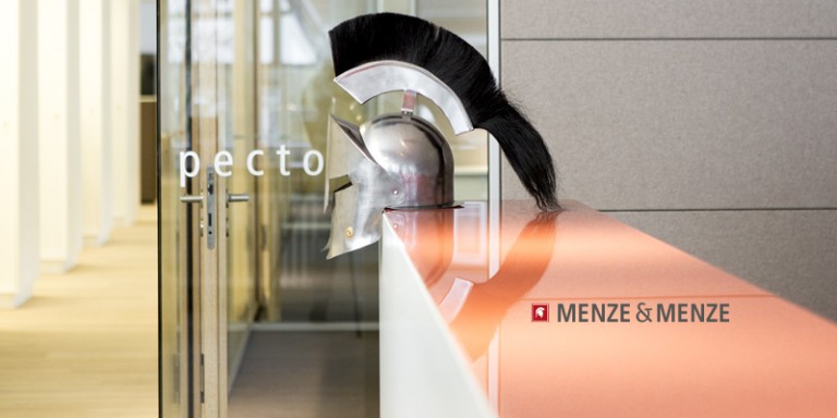 Ein Ritterhelm, der seitlich fotografiert wurde und dieser liegt auf einem orangenen Tresen. Dahinter ist links ein Flur, in der Mitte eine Glaswand, die einen Raum andeutet und rechts eine graue Wand. Unten rechts ist das Menze & Menze Logo