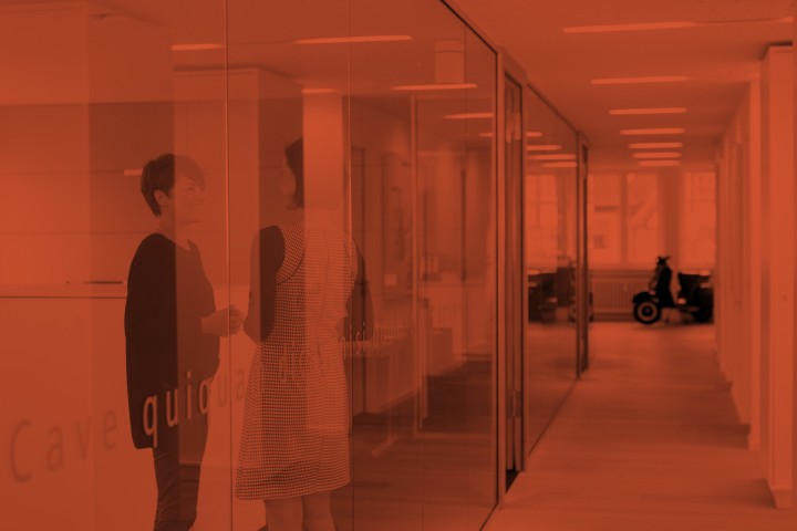 Gespräch zwischen Mitarbeiterinnen, die hinter einer Glaswand stehen. Das Bild hat einen roten Filter