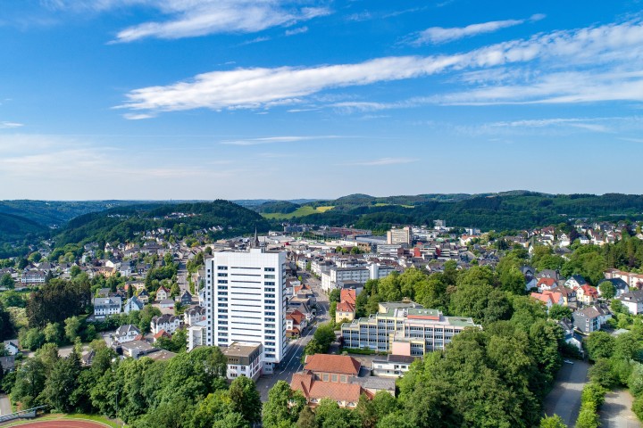 Ausblick über die Stadt Gummersbach. Zu sehen sind viele Gebäude, im Hintergrund grüne Wälder und Felder unter blauem Himmel.
