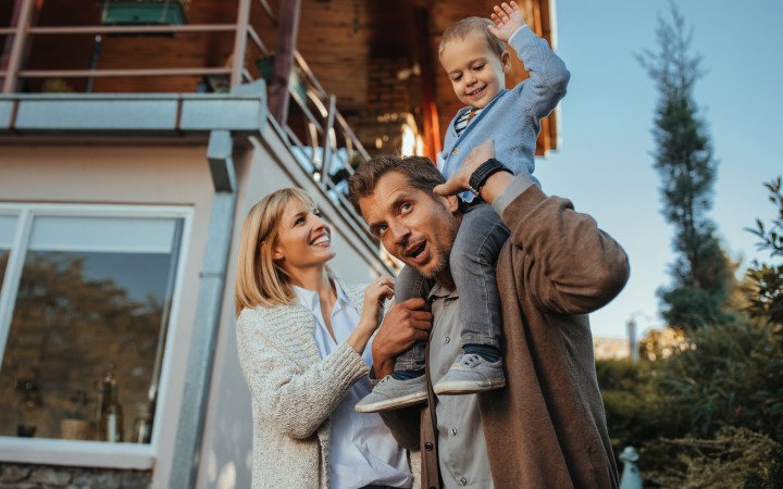 Glücklich lachende Familie vor ihrem Haus und der Vater trägt den kleinen blonden Sohn auf den Schultern