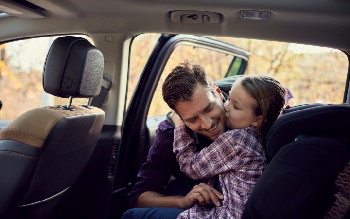 Vater setzt seine kleine Tochter in den Kindersitz auf der Rückbank eines Autos, während diese ihm einen Kuss auf die Wange gibt, beide tragen karierte Hemden