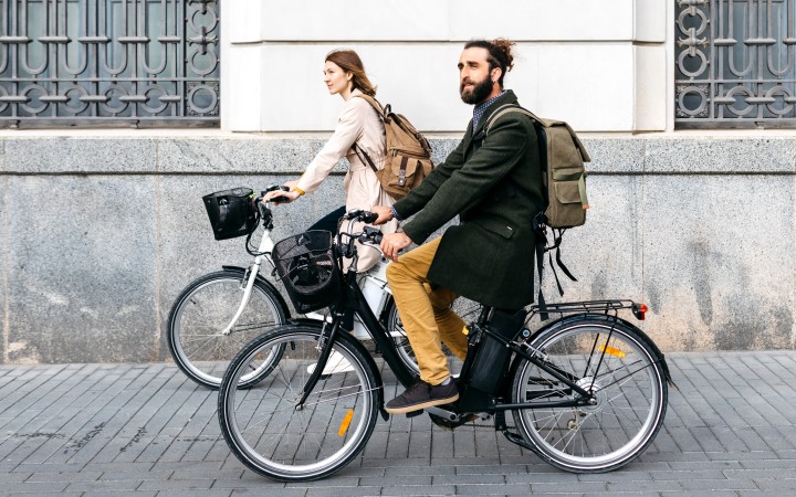Mann und Frau fahren nebeneinander auf E-Bikes auf der Straße an einer Häuserreihe vorbei