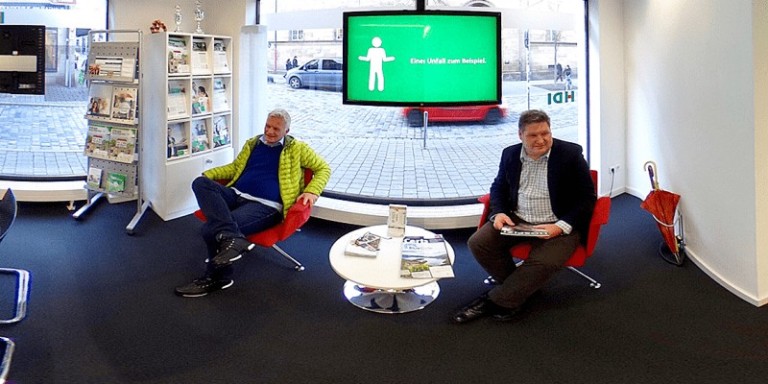 Man sieht die Agentur von innen. Der Fokus liegt auf einer kleinen Sitzgruppe, die aus einen Beistelltisch und zwei Stühlen besteht, auf denen zwei Männer sitzen. Dahinter hängt ein Fernseher vor der Fensterscheibe. Links neben dem Fenster steht eiMan sieht die Agentur von innen. Der Fokus liegt auf einer kleinen Sitzgruppe, die aus einen Beistelltisch und zwei Stühlen besteht, auf denen zwei Männer sitzen. Dahinter hängt ein Fernseher vor der Fensterscheibe. Links neben dem Fenster steht ein Regal und ein Aufsteller mit Infomaterial von HDI.n Regal und ein Aufsteller mit Infomaterial von HDI.