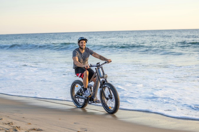 Junger Mann mit Helm fährt lachend auch einem E-Bike am Strand entlang