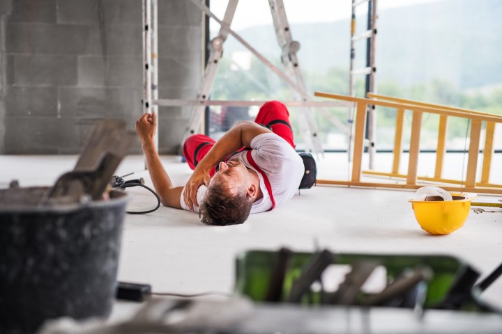 Mann in Arbeitshose leigt auf Boden. Man sieht eine umgekippte Leiter, neben ihm liegt ein gelber Schutzhelm, im Hintergrund ist eine weitere Leiter zu erkennen und ein Gerüst, im Vordergrund stehen andere Arbeitsutensilien
