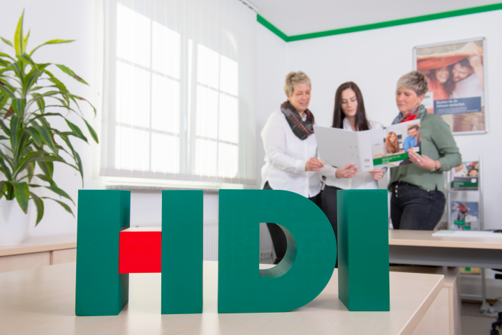 HDI Logo ist im Vordergrund, dahinter stehen drei Mitarbeiter hinter einem Tisch und gucken in ein Ordner hinein