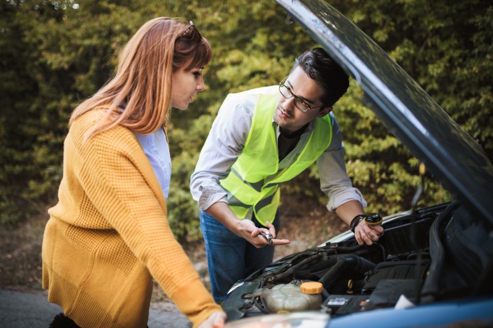 Eine Frau und ein Mann, der eine Warnweste an hat und hat etwas in der Hand. Beide gucken in die Motorhaube eines Autos.