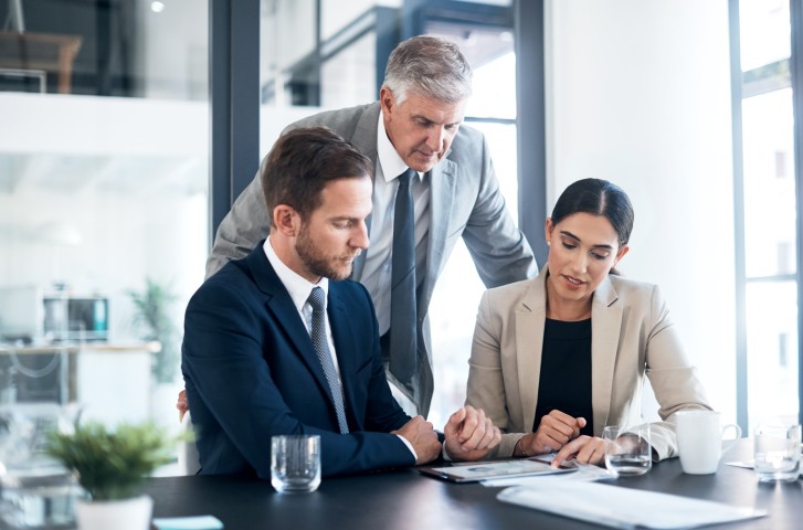 Zwei Männer mittleren Alters und eine Frau unterhalten sich geschäftlich. Alle drei haben einen Anzug an, die beiden Männer mit Krawatte. Der linke Mann hat braunde Haare und einen Bart, der Mann in der Mitte hat graues Haar und kein Bart. Die Frau links ist geschminkt und trägt einen Zopf mit langen schwarzen Haaren.