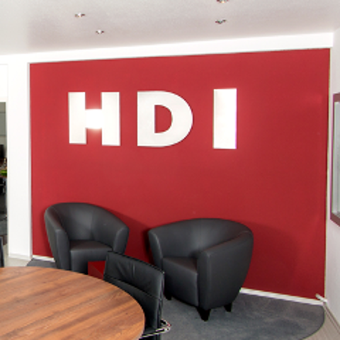 Agentur von innen, in der linken unten Ecke steht ein brauner Tisch mit einem schwarzen Stuhl, in de Mitte an der roten Wand un dem silbernen HDI Logo hinten stehen zwei schwarze Sessel