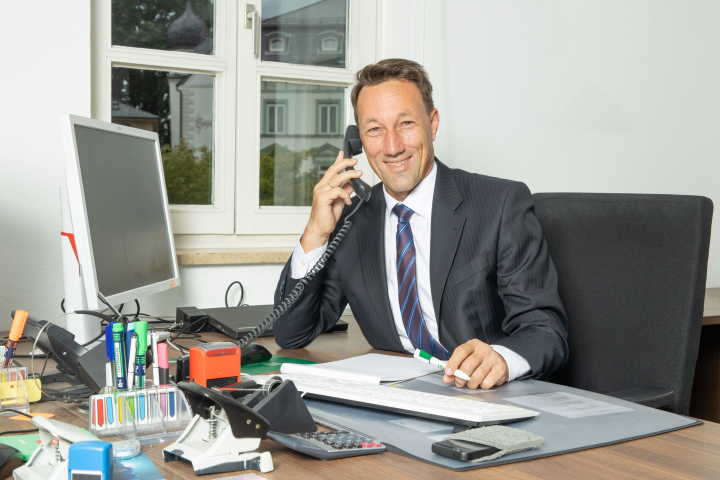 Alexander Kastenhuber sitzt an einem Schreibtisch. Auf diesem Schreibtisch liegen Locher, Tasschenrechner, Stifte, Handy, telfon und PC. Er ist am telefonieren und hält einen Stift in der rechten Hand. hinter Ihm ist ein Fenster, dort steht ein weißes Haus und grüne Bäume