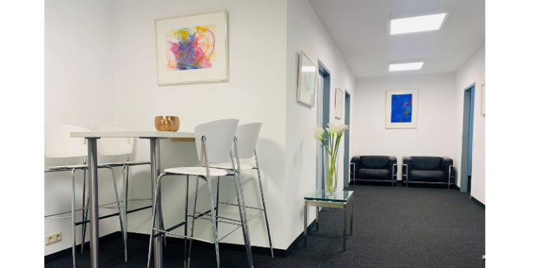 Man sieht einen hohen Tisch mit vier Stühlen. Daneben schaut man einen kleinen Gang hinunter von dem mehrere Türen abgehen. Am Ende stehen zwei Sessel. Die Wände sind mit Bildern geschmückt.