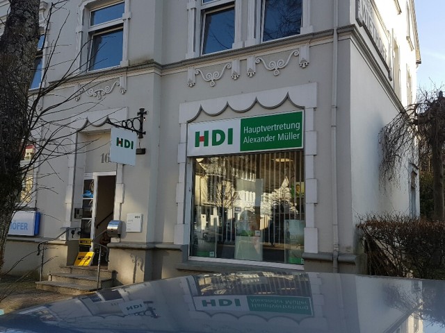 Man sieht das Agenturgebäude von außen. Es ist ein freistehendes Haus. Man sieht eine Tür und ein Schaufenster. Über dem Fenster hängt ein Schild mit dem HDI Logo und dem Namen der Agentur.