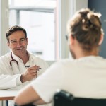 Auf der einen Seite mit dem Bilck zum Patienten sitzt ein Arzt mit einem Stift in der Hand und gegenüber von dem Arzt mit dem Rücken zur Kamera sitzt der Patient im Rollstuhl. 