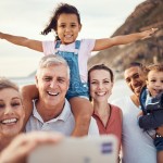 Auf dem Bild sinf zwei ältere Menschen, zwei Jüngere Menschen und zwei Kinder. DIe ältere Frau hält das handy, um ein Foto zu machen. Das Mädchen sietzt au den Schultern von dem älteren Mann und neben ihm steht eine Frau und neben der Frau steht ein Mann, der einen Jungen auf dem Arm hält. die Familie steht vor dem wass und einem Berg. 