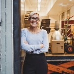 Eine Frau ist links an einem Türrahmen angelehnt und rechts hinter hier sieht man Umzugkartons, Stühle und eine Lampe. 