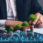 Mann in Anzug, vor ihm auf den Tisch sind mehrere Stapel aus Münzen, auf der obersten Münze jeden Stapels ist ein kleiner Baum, außerdem sind verschiedene Grafiken, Kurven  und Zahlenwerte zu sehen