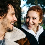 Links ist ein Mann und rechts eine Frau, sie lehnen sich beide an einen Baum und lächeln sich gegenseitig an. 