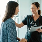 Links ist eine Patienten, die auf einer Liege sitzt und auf der rechten Seite steht eine Ärztin mit grünen Kittel. In der linken Hand hält sie ein Brett mit Zettel und mit die rechte Hand liegt auf der Schulter, der Patienten. 