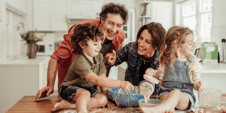 Eine Familie, die Eltern stehen hintder dem Tisch und zwei Kinder sitzen auf dem Tisch. In der Mitte der Familie sthet eine Schüssel mit Teig.