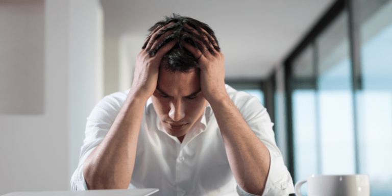Der Mann hat seine Ellenbogen auf den Tisch gestellt und fässt sich mit den Händen an seinen Kopf. Sein Gesicht richtet sich nach unten. 