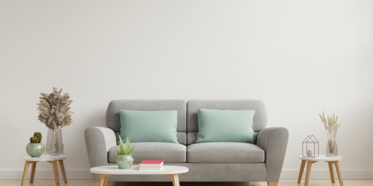 Das Sofa ist hellgrau und vor dem Sofa steht ein kleiner weißer Tisch, auf dem eine Pflanze und ein Buch drauf liegen.