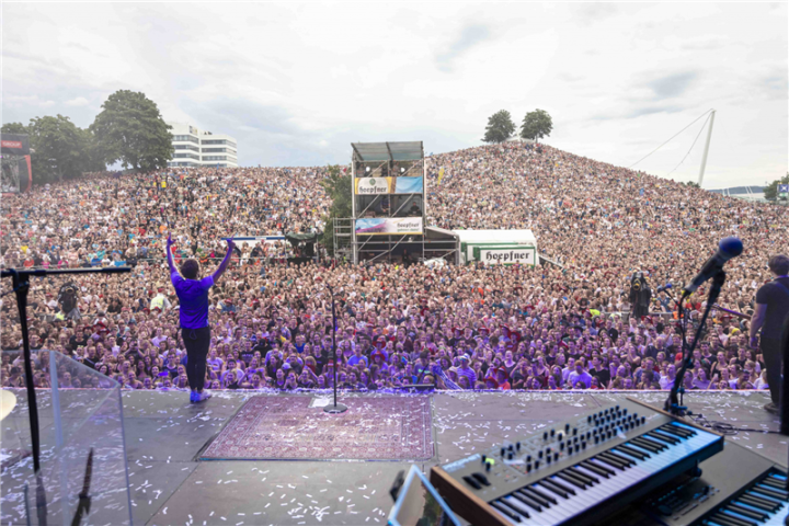 Das Foto wurde von einer Bühne gemacht. Links Auf der Bühner steht der Sänger und Rechts steht ein Klavier. Vor der Bühne stehen ganz viel Menschen und Mitten in der Menge steht ein Gerüst. 