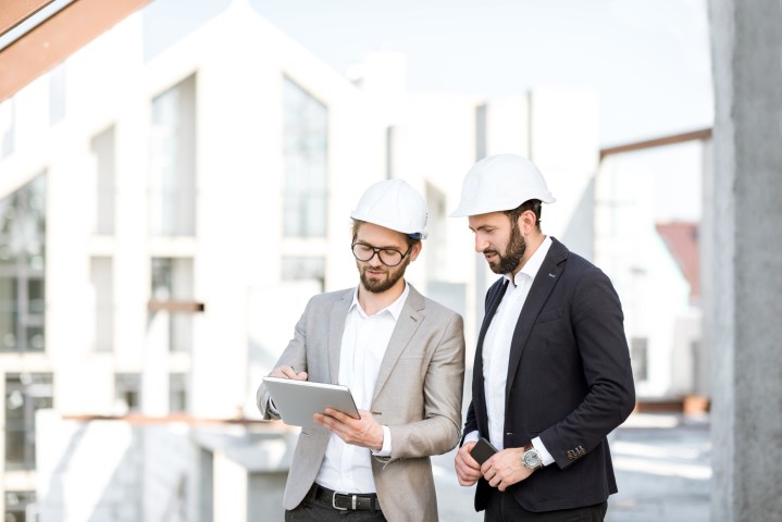 Zwei Architekten auf einem Bauobjekt, die sich unterhalten. Beide Herren tragen einen weißen Bauhelm und einen Anzug.