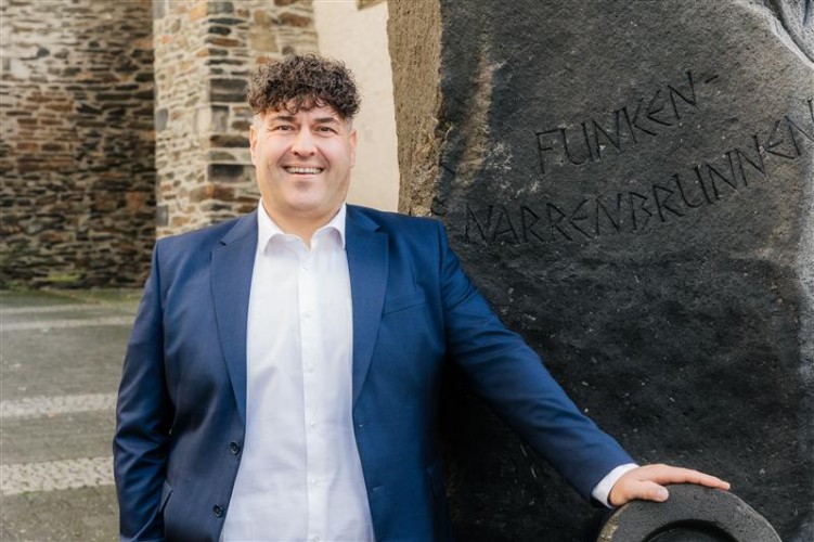 Schneider lächelt in die Kamera und hinter ihm ist ein Stein mit der Aufschrift "Funken-Narrenbrunnen"