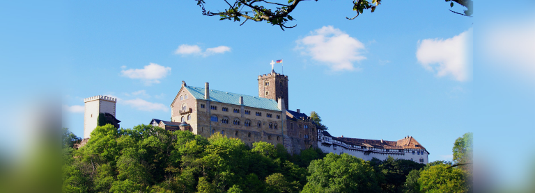 Altes burg-ähnliches Gebäude, Bäume im Vordergrund, der Himmel ist blau und die Sonne scheint