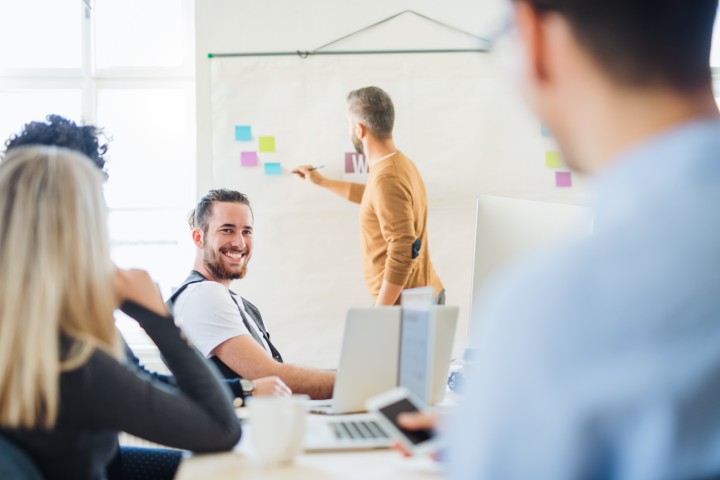 Mehrere Personen sitzen an Besprechungstisch auf dem Laptops und eine Kaffeetasse stehen, dahinter steht ein Mann der gerade etwas auf ein Whiteboard zeichnet