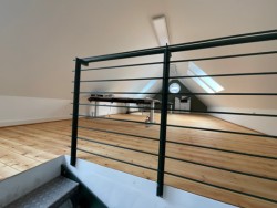 Zusehen ist das Dachgeschoss der Agentur Rauh. Ein hellbrauner Parkettboden und das Schwarze Gelände der Treppe.