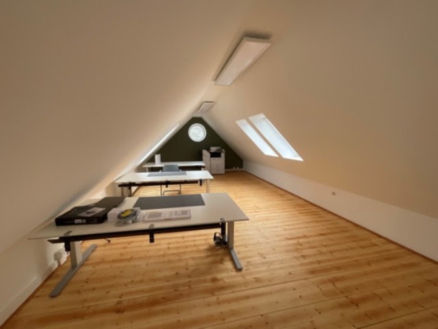 Zusehen ist das Dachgeschoss der Agentur Rauh. Ein hellbrauner Parkettboden und  drei große Schreibtische auf der Linken Seite, zwei Dachfenster auf der echten Seite.