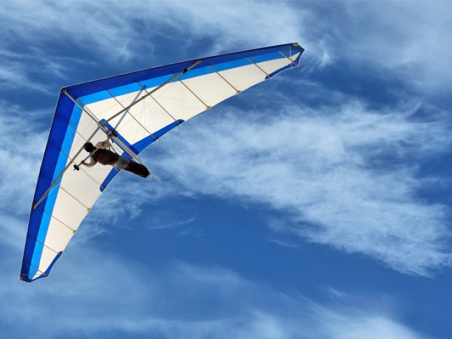 Blau-weißer Drachenflieger in dem eine Person fliegt. Der Himmel ist blau und leicht bewölkt.