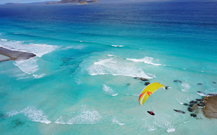 Gelber Gleitschirmflieger über blauem Wasser. Im Wasser sind einige Felsen zu sehen, im Hintergrund sieht den Horizont, ein paar kleine Inseln und den teils wolkenbedeckten Himmel.