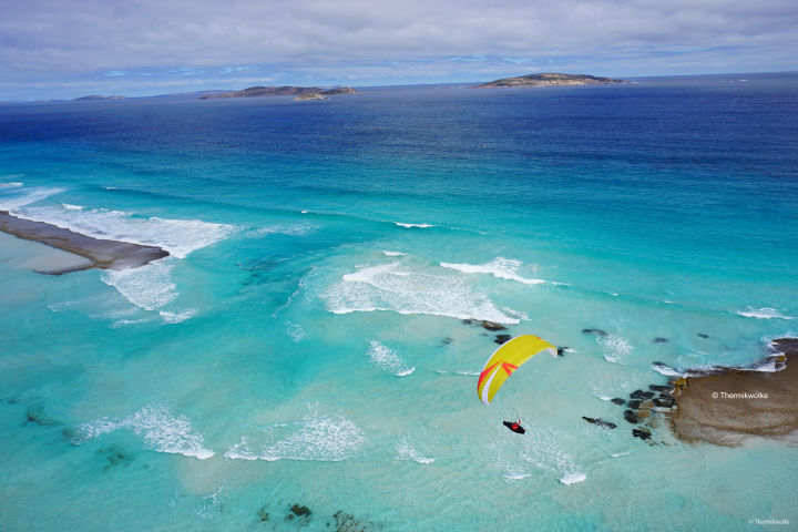 Gelber Gleitschirmflieger über blauem Wasser. Im Wasser sind einige Felsen zu sehen, im Hintergrund sieht den Horizont, ein paar kleine Inseln und den teils wolkenbedeckten Himmel.