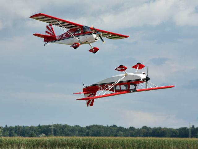 Zwei rot-weiße Modellflugzeuge in der Luft. Sie fliegen über ein Feld. Im Hintergrund sieht man eine Waldkante. Der Himmel ist leicht wolkenverhangen. Eines der Flugzeuge steht auf dem Kopf.