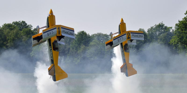 Zwei Modellflieger in gelb die senkrecht nach oben fliegen. Beide ziehen eine weiße Auspuffwolke hinter sich her. Im Hintergrund sieht man Bäume.